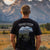 A man faces the Teton mountains wearing a black shirt with a stylized grizzly bear on the back. Within the shape of the bear is a photo from the Snake River Overlook. 